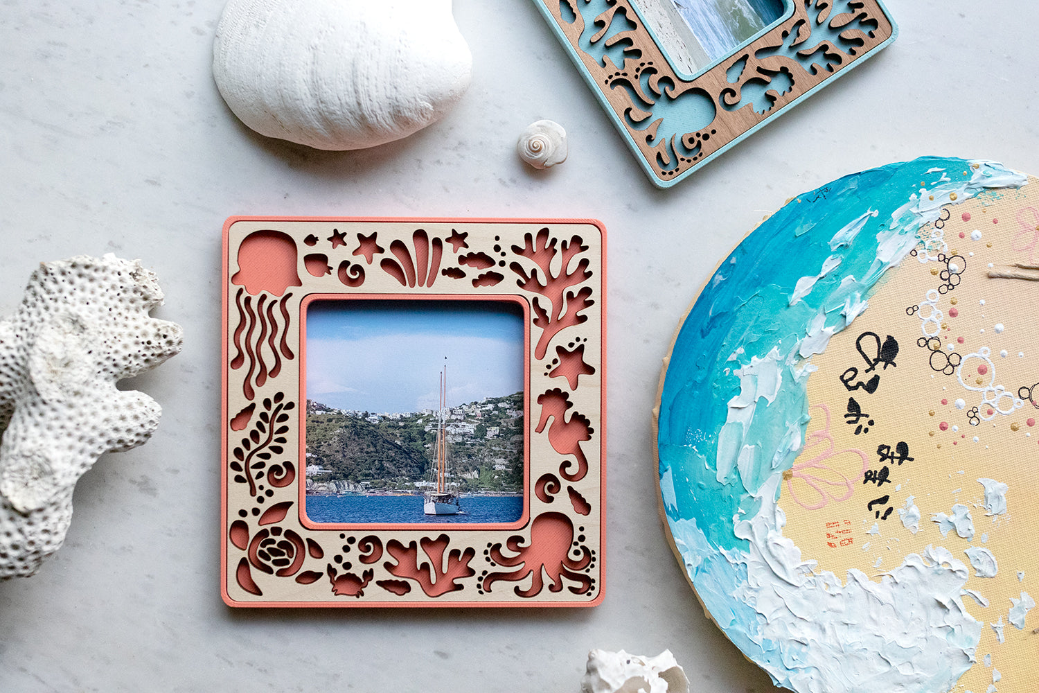 A standard polaroid frame with sea creatures design. Frame is coral with laser cut maple inlay. The frame is displayed next to a painting of the ocean and seashells from the beach. Customization options make a great beach house gift.