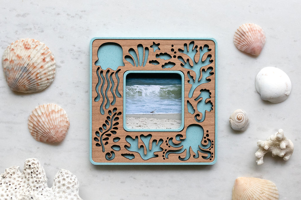 Decorative blue Polaroid Go magnetic frame with laser cut ocean creatures on a cherry wood inlay. The frame is lying on a white surface with seashells all around and displaying a photo of the ocean.