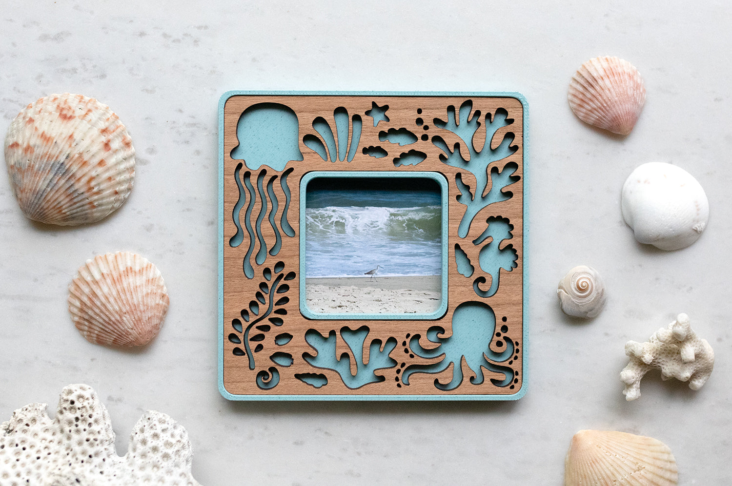 Decorative blue Polaroid Go magnetic frame with laser cut ocean creatures on a cherry wood inlay. The frame is lying on a white surface with seashells all around and displaying a photo of the ocean.
