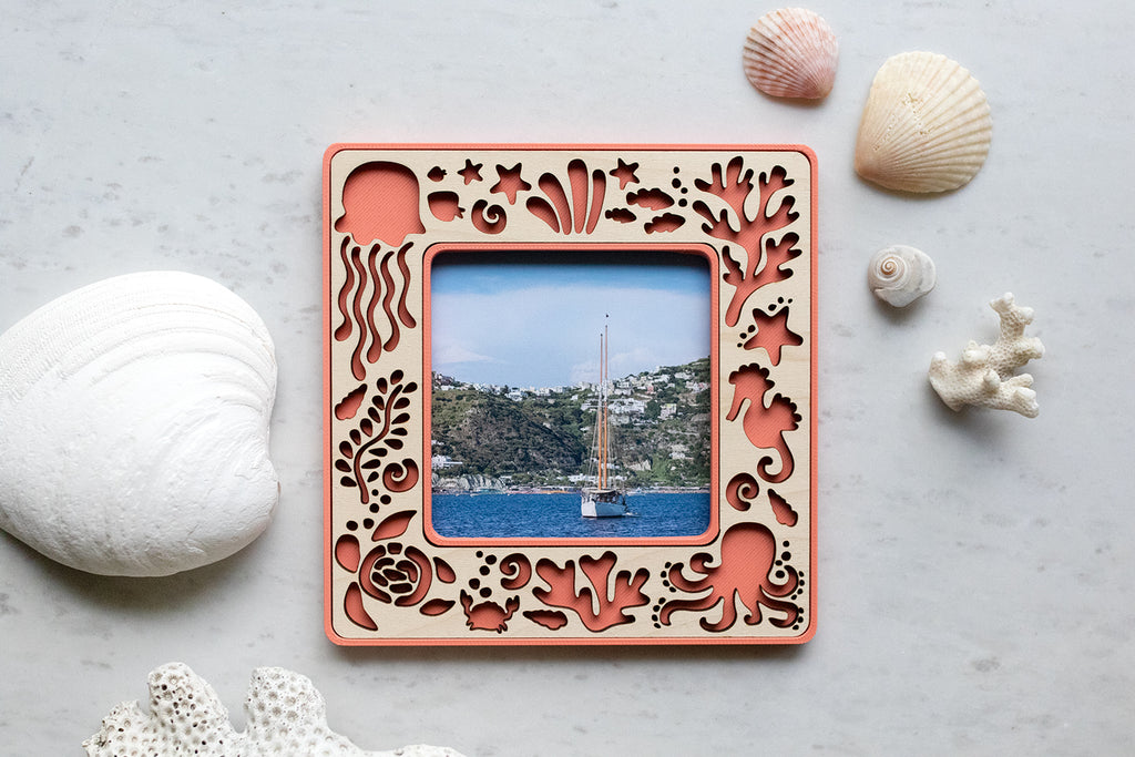 A small magnetic coral beach frame with sea creatures, including: an octopus, a sea turtle, jellyfish, starfish, and a crab. The magnetic frame has a laser cut maple inlay and is surrounded by seashells.
