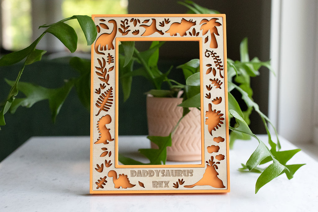 A laser cut dinosaur wooden picture frame keepsake with orange 3D printed housing and maple wood inlay. The frame is decorated with laser-cut dinosaur and prehistoric plant designs. The text "DADDYSAURUS REX" is at the bottom. The frame is displayed in front of a green plant.