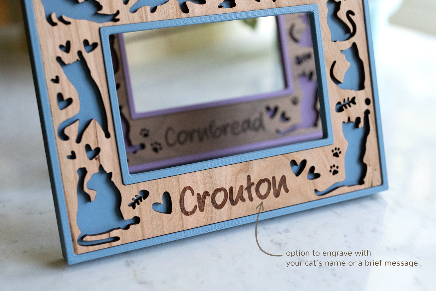 A custom blue cat frame that has the "option to engrave with your cat's name or a brief message". The pictured frame is Fujifilm Instax Wide size and includes the laser engraved name "Crouton". The cat frame in the background is purple with walnut, and the name "Cornbread" is engraved on the wooden inlay.