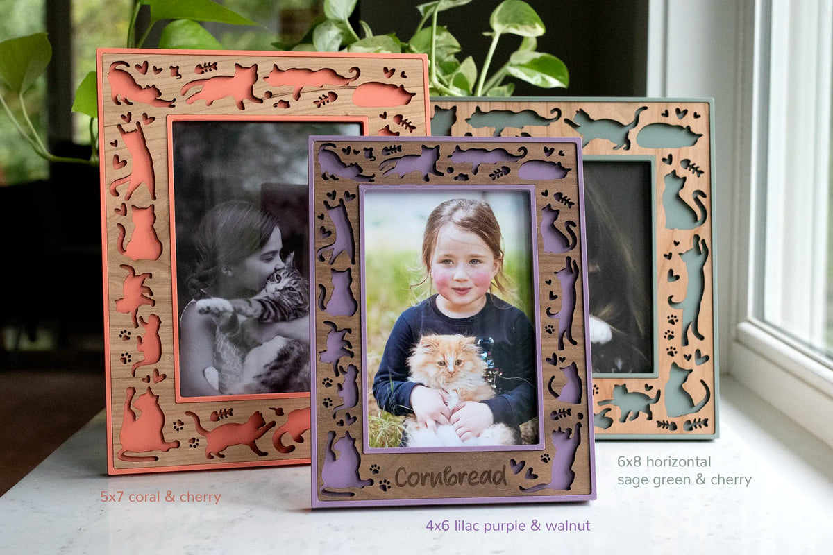 Three personalized cat picture frames in different sizes and colors, with laser-cut wood borders of cat silhouettes, paw prints, and hearts: a 5x7 coral and cherry frame, a 4x6 lilac purple and walnut frame engraved with the pet name "Cornbread," and a 6x8 horizontal sage green and cherry pet photo frame.
