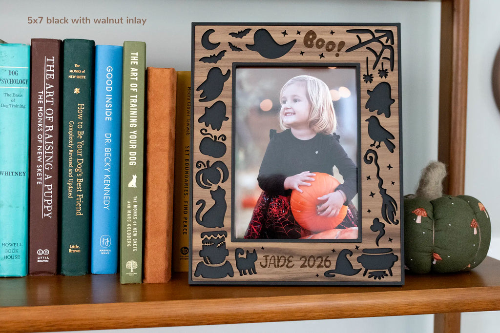Personalized Halloween picture frame, 5x7 in black with walnut inlay, with laser-cut ghosts, bats, black cat, and pumpkins, engraved "Jade 2026," on a bookshelf