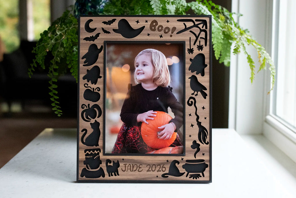 Personalized Halloween picture frame in black and walnut with laser-cut ghosts, bats, pumpkins, broom, and "Boo!", engraved "Jade 2026," on a white table by a window.