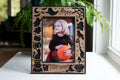Personalized Halloween picture frame in black and walnut with laser-cut ghosts, bats, pumpkins, broom, and "Boo!", engraved "Jade 2026," on a white table by a window.