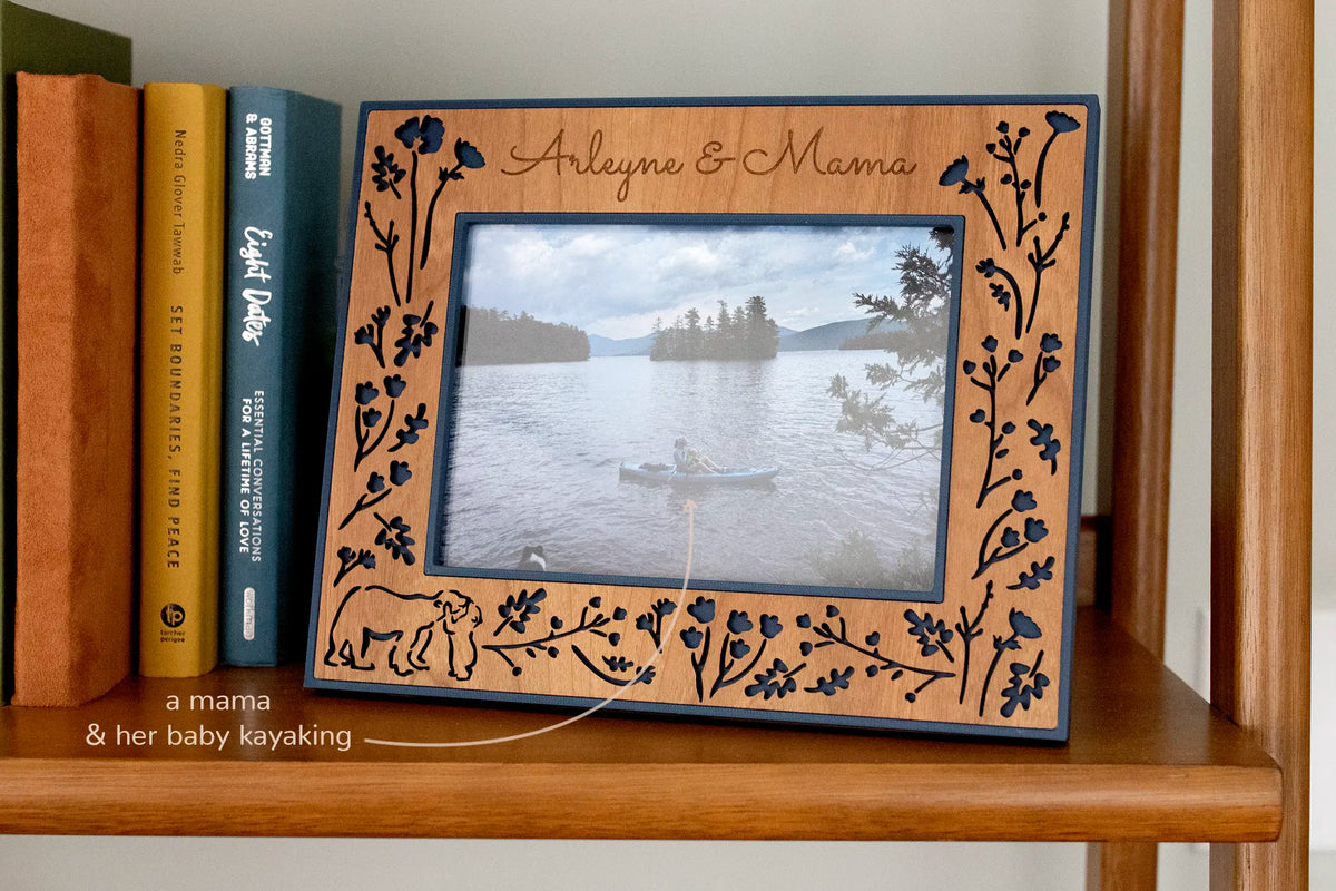 A 5x7 horizontal Mama and Baby Bear picture frame on a bookshelf, engraved with “Arleyne & Mama.” The cherry wood inlay is laser-cut with wildflowers and a mama bear and cub over a dark blue housing, framing a photo of a mother and baby kayaking on a mountain lake.
