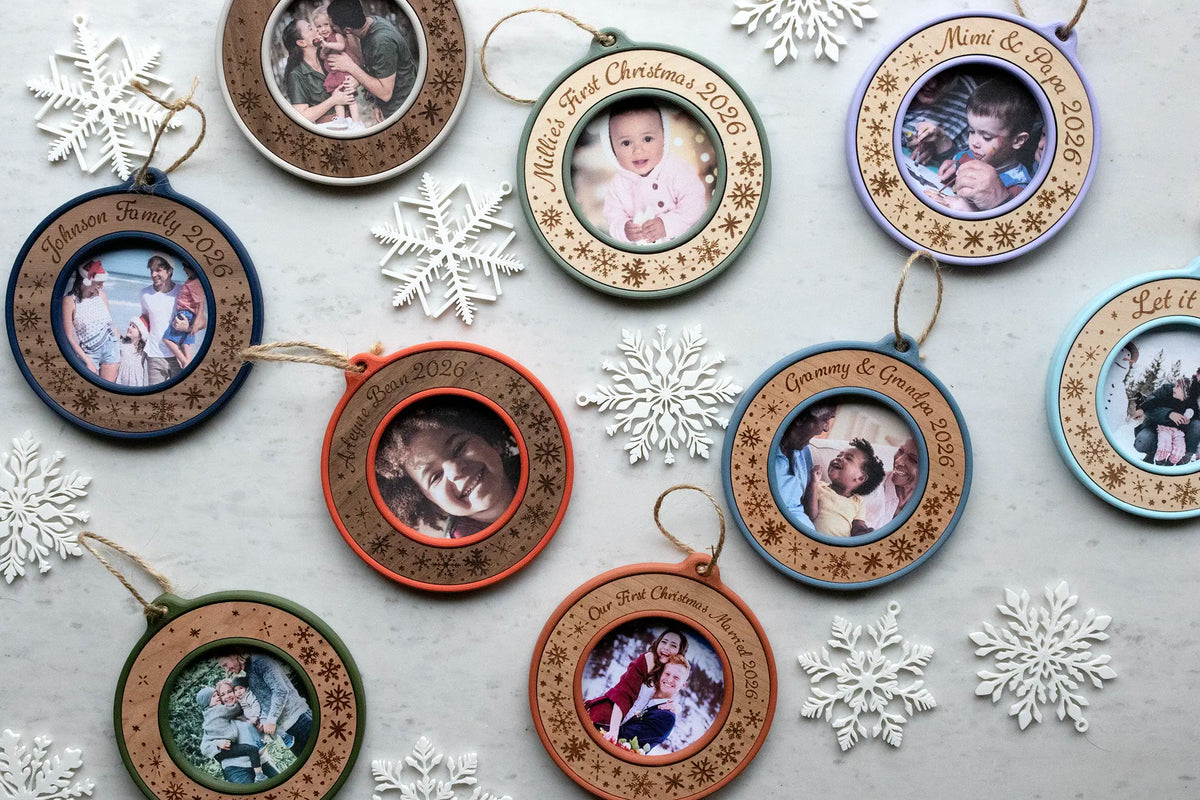Flat lay of nine personalized Christmas photo ornaments in nine housing colors with maple, cherry, and walnut inlays, each a mini picture frame laser-engraved with snowflakes and custom text such as "Johnson Family 2026," "Millie's First Christmas 2026," and "Grammy & Grandpa 2026." They're arranged on white marble among white snowflake decorations.
