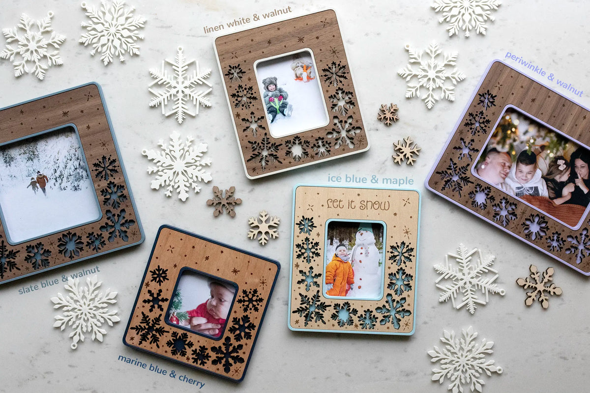 Five laser-cut snowflake frames with magnets for winter fridge decor. These snowflake frames are available in 4 sizes: Instax Mini, Instax Wide, Standard Polaroid, and Polaroid Go. They are surrounded by snowflake ornaments and lying on a white surface.