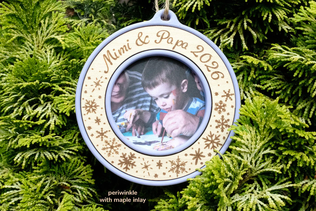 Grandparent photo ornament in periwinkle purple with a maple inlay, laser-engraved "Mimi & Papa 2026" with snowflakes, holding a photo of a young boy painting with his grandparents, hanging on evergreen branches.