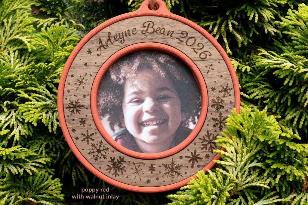 Custom name Christmas ornament in poppy red with a walnut inlay, laser-engraved "Arleyne Bean 2026" with snowflakes, a round mini picture frame holding a photo of a smiling girl with curly hair.