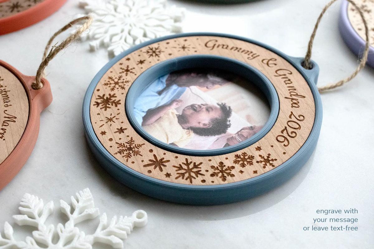 Angled view of a slate blue and cherry grandparents photo ornament engraved "Grammy & Grandpa 2026," showing the depth of the 3D-printed mini picture frame and its laser-engraved snowflakes. Caption text reads "engrave with your message or leave text-free."
