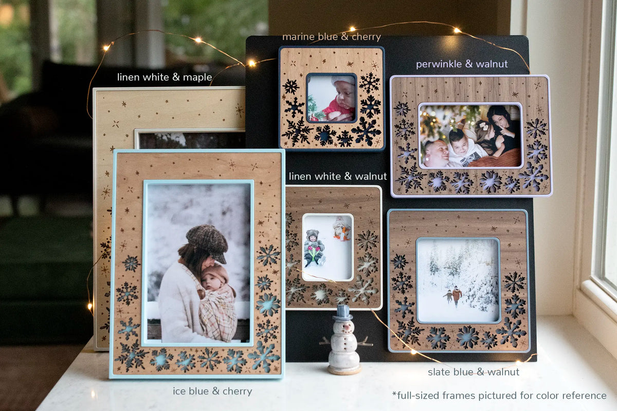 An assortment of laser-cut snowflake Christmas keepsake frames in many colors and woods — linen white, marine blue, periwinkle, ice blue, and slate blue, in maple, cherry, and walnut — styled with fairy lights and a ceramic snowman, holding family Christmas photos. These winter holiday frames are hand-sanded, finished, and assembled in Western North Carolina. Magnetic Instax Mini, Instax Wide, and Polaroid sizes hang on a black magnetic board.