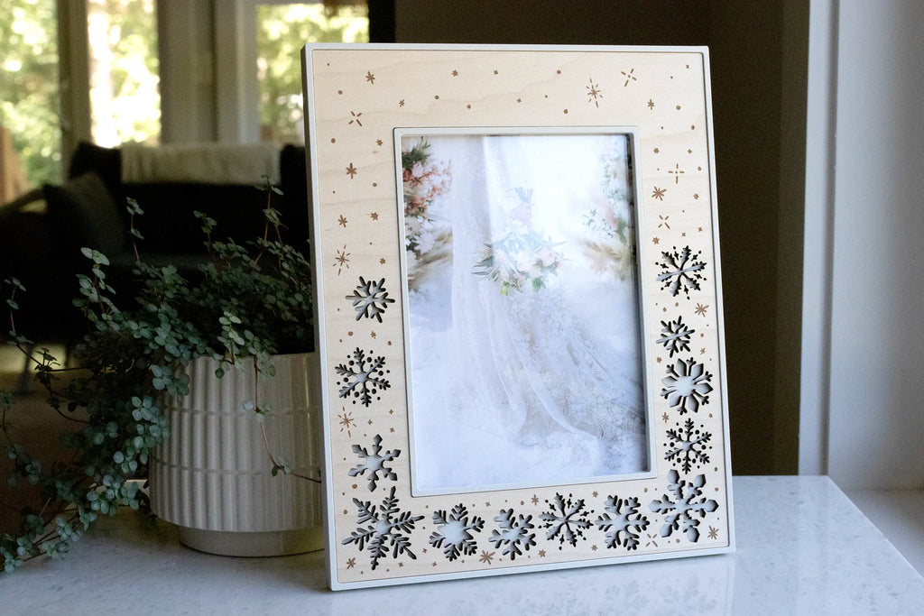 A linen white and maple snowflake picture frame holding a photo of a bride with her bouquet in the snow, displayed on a marble counter beside eucalyptus. This laser-cut winter wedding keepsake features falling snowflakes and engraved snow sparkles in a wood inlay.
