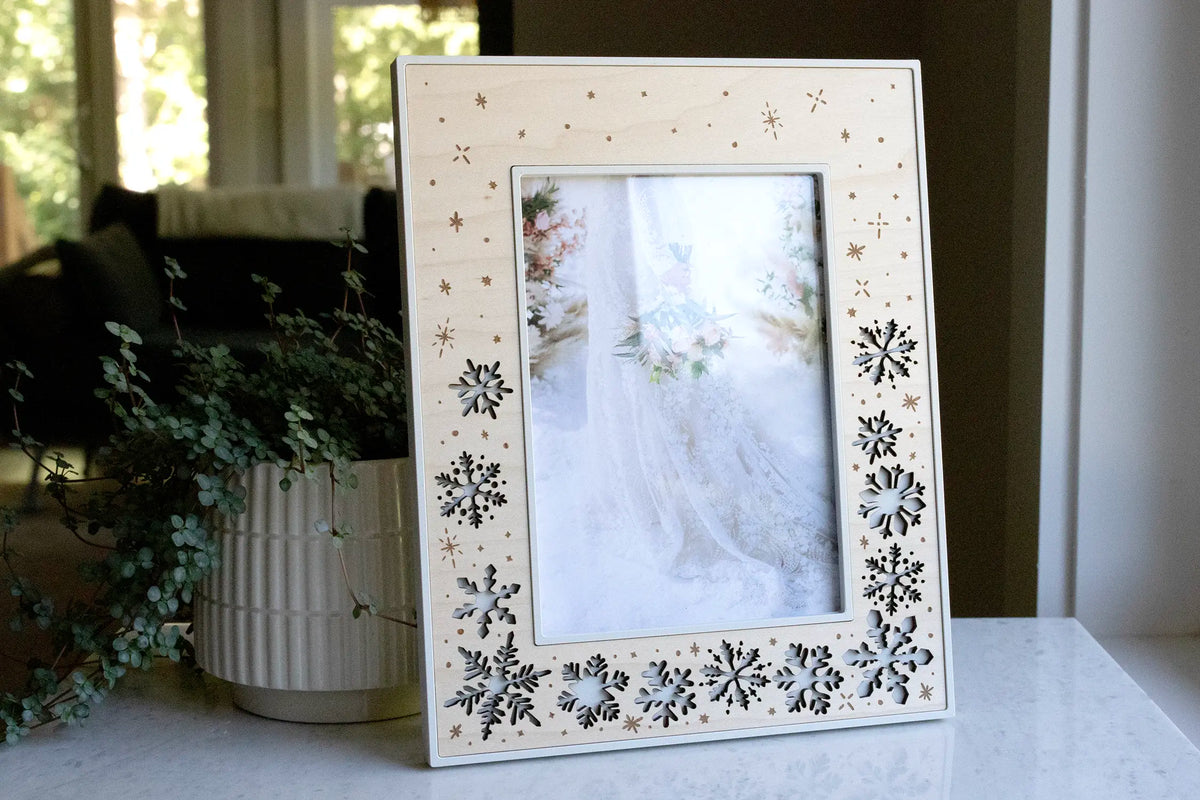 A linen white and maple snowflake picture frame holding a photo of a bride with her bouquet in the snow, displayed on a marble counter beside eucalyptus. This laser-cut winter wedding keepsake features falling snowflakes and engraved snow sparkles in a wood inlay.