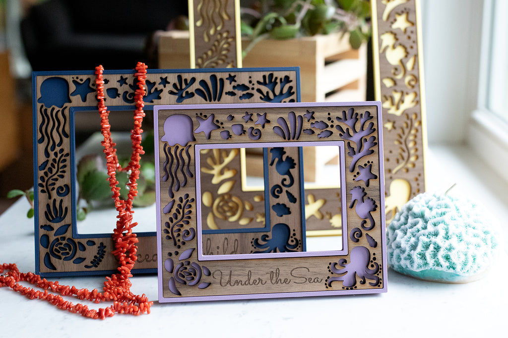 Ocean lover laser cut picture frames on a white counter with a coral necklace and blue coral candle. The picture frames are colorful shown in yellow, blue, and purple and they include whimsical sea creature designs in the wood.