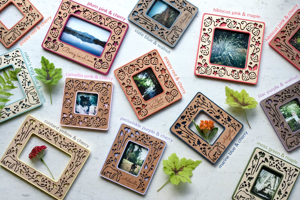 An assortment of colorful, laser cut strawberry and bears Polaroid frames. The frames are lying on a white surface, surrounded by flowers and leaves. They have an intricate, feminine design and real wooden inlays. Some of the frames include an engraved personalized message.