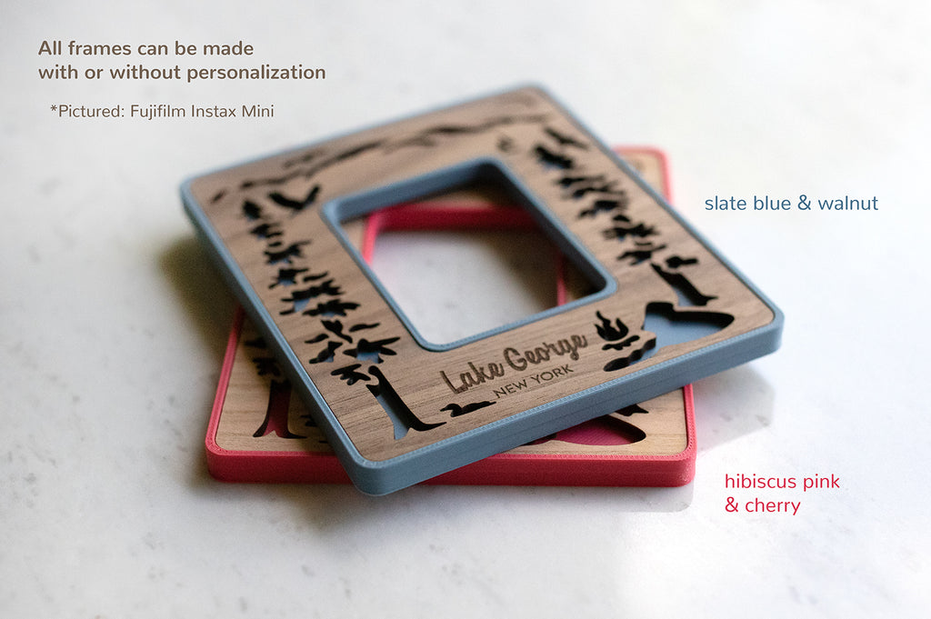 Two Instax Mini magnetic Polaroid photo frames stacked on top of each other and lying on a white counter. The top left corner of the image displays the words: "All frames can be made with or without personalization. Pictured: Fujifilm Instax Mini". The top frame in walnut and slate blue is personalized with the words "Lake George New York."