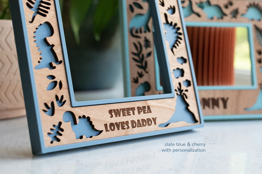 Close up detail view of a wooden frame with laser cut dinosaurs and a message that reads "Sweet Pea Loves Daddy". The frame is blue with cherry wood border. Fully customizable with a laser-engraved message and available in a variety of wood types and frame colors.
