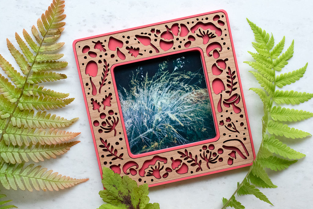 A bright pink magnetic frame with mushroom design. The frame holds a standard size polaroid photo. The frame has a detailed cherry inlay with laser cut mushrooms and acorns. The mushrooms include amanita mushrooms, porcini mushrooms, and other common mushroom types.