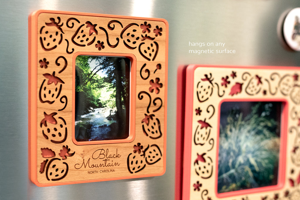 A close-up detail view of an Instax Mini frame and a Standard Polaroid frame on the front of a stainless steel refrigerator. The frames have an intricate laser cut design, displaying strawberries and strawberry flowers. The words "Black Mountain North Carolina" are engraved in the wooden inlay. The frames are red with cherry wood and pink with birch wood.