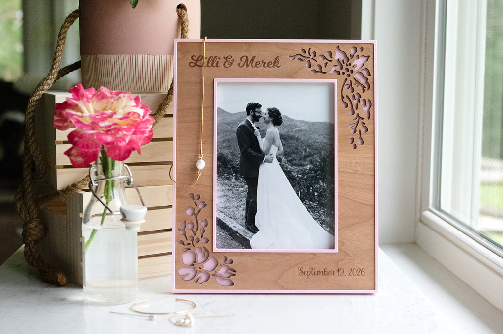 Pink wedding flower picture frame with laser cut cherry wood inlay. The frame is custom engraved with the names "Lilli & Merek" and wedding date "September 19, 2026". There is a delicate pearl bridal wedding necklace, earrings, and a bright pink flower next to the frame.