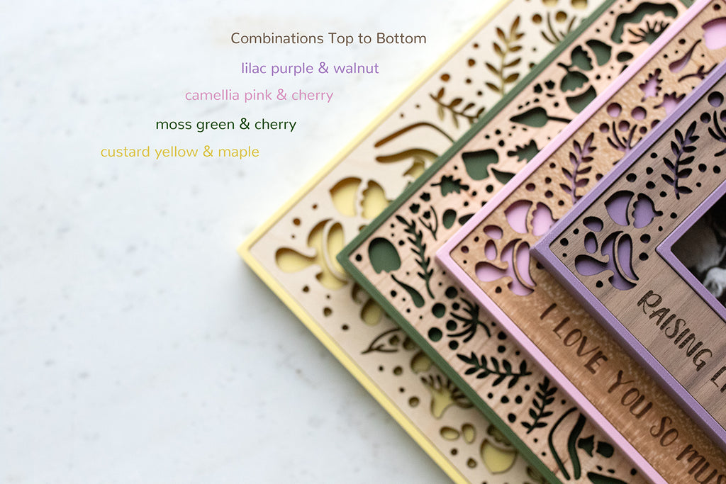 Wooden frames with laser-cut mushroom and acorn designs in various color combinations, including lilac purple with walnut, camellia pink with cherry, moss green with cherry, and custard yellow with maple. The text on the frames read "I love you so mush" and "raising little fun-gis."