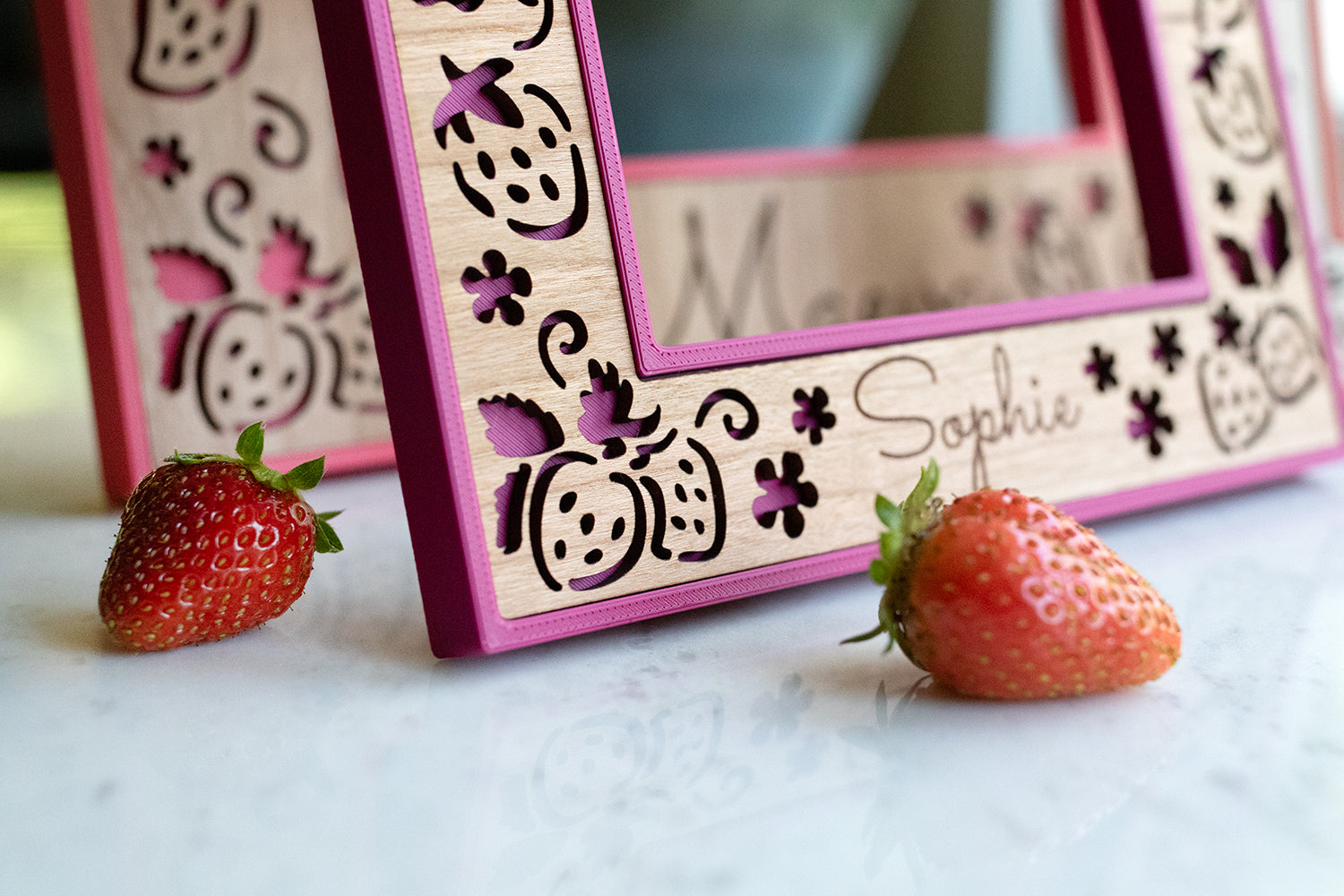 A close up, detail view of a laser cut strawberry frame in a vibrant color called "plum pink" with a cherry wood inlay. Real strawberries lay on a white surface in the foreground.
