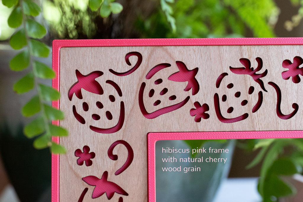 A close up detail front view of a laser cut strawberry picture frame. The image reads "hibiscus pink frame with natural cherry wood grain". The frame is in natural light, and there is greenery in the background.