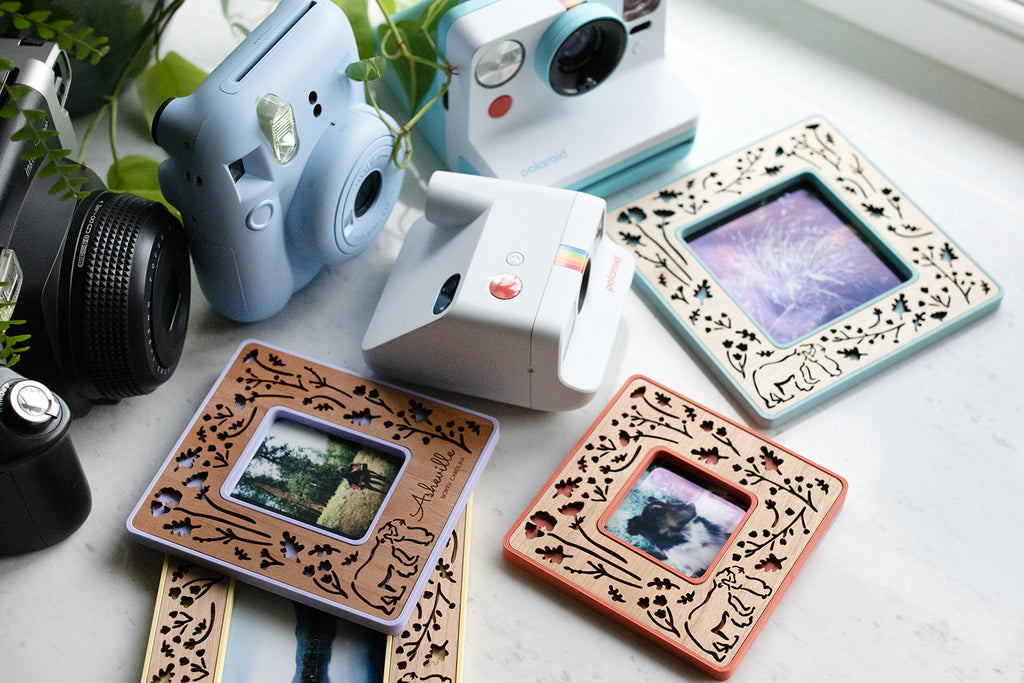 An assortment of four Polaroid picture frames lying on a white table next to Polaroid cameras and Fujifilm Instax cameras. The frames are displaying images of dogs, plants, and mountain scenery.