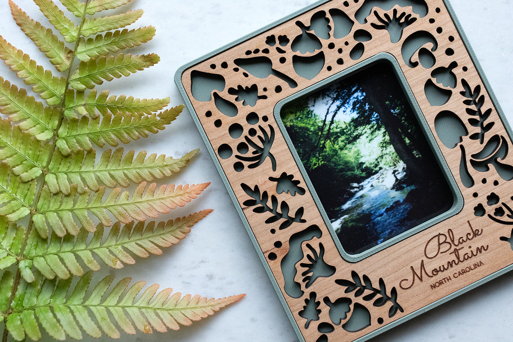 A close-up detail view of a whimsical mushroom magnetic frame lying next to a large green fern frond. The frame features a delicate pattern of various mushroom types and acorns, including amanita mushrooms, porcini mushrooms, and the classic toadstool mushroom design.