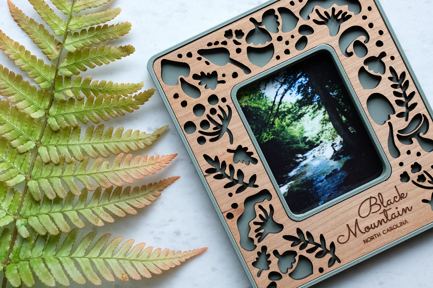 A close-up detail view of a whimsical mushroom magnetic frame lying next to a large green fern frond. The frame features a delicate pattern of various mushroom types and acorns, including amanita mushrooms, porcini mushrooms, and the classic toadstool mushroom design.