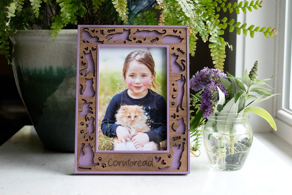 Personalized cat picture frame in lilac purple with a laser-cut walnut wood border of cat silhouettes, paw prints, and hearts, engraved with the pet name "Cornbread," holding a photo of a girl with her fluffy orange kitten, next to fern and lavender plants.