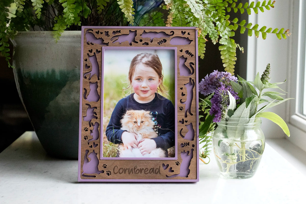 Personalized cat picture frame in lilac purple with a laser-cut walnut wood border of cat silhouettes, paw prints, and hearts, engraved with the pet name "Cornbread," holding a photo of a girl with her fluffy orange kitten, next to fern and lavender plants.