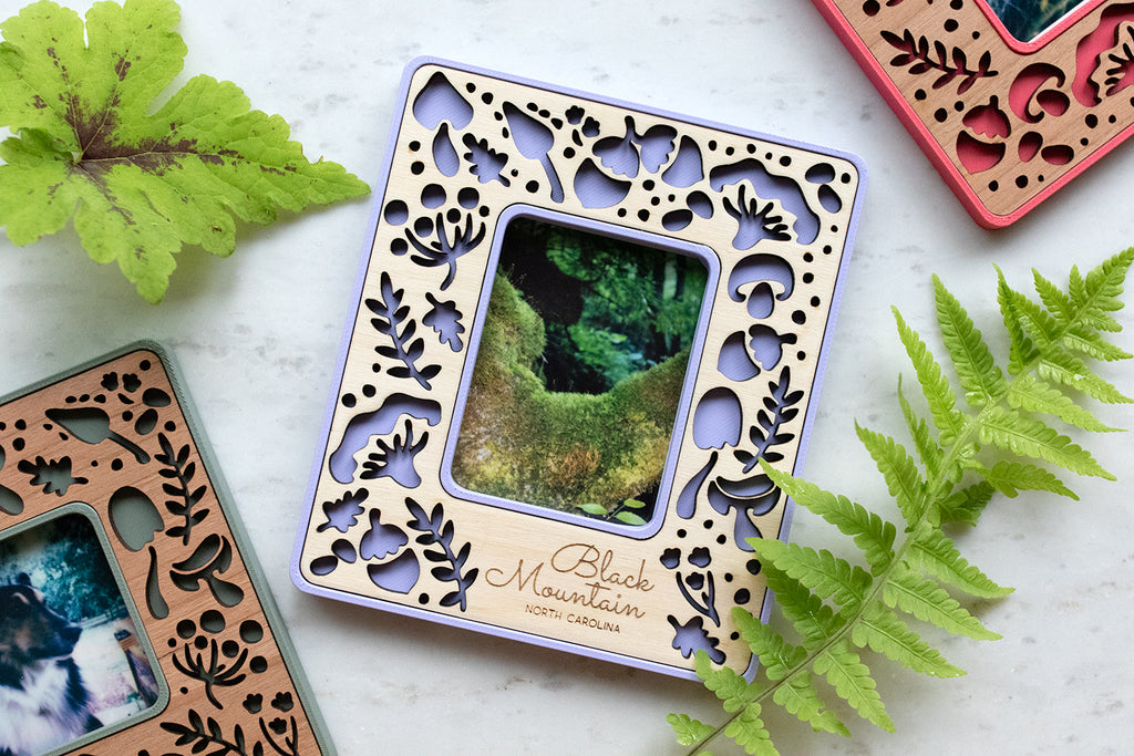 A pretty, light purple magnetic picture frame featuring whimsical mushrooms. Frames can be personalized and are available in various wood types and colors. Three mushroom frames are displayed in periwinkle purple, sage green, and a bright hibiscus pink color. The frames are lying on a white surface and are surrounded by greenery.