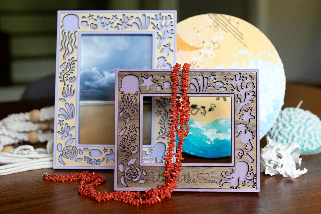 Two "Under the Sea" themed laser cut picture frames shown with a white seashell, coral necklace, coral candle, and ocean art. The frames are two shades of purple and include both walnut and birch inlay options.