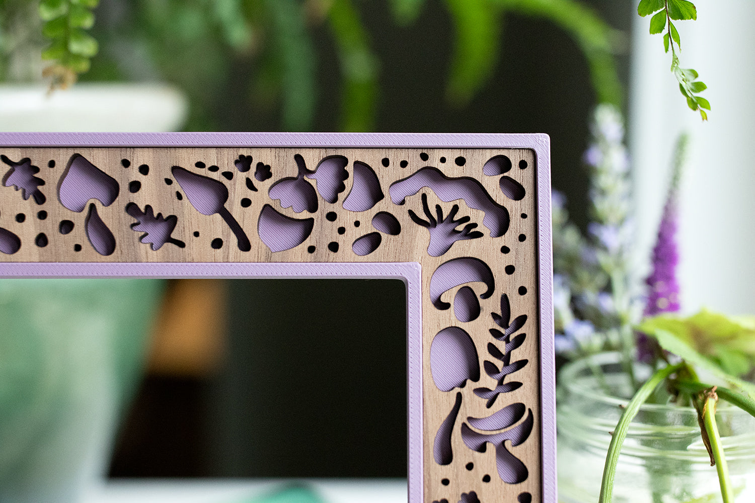 A close-up, detail view of a beautiful laser cut mushroom frame with purple housing and a walnut inlay. There are purple flowers and greenery in the background.
