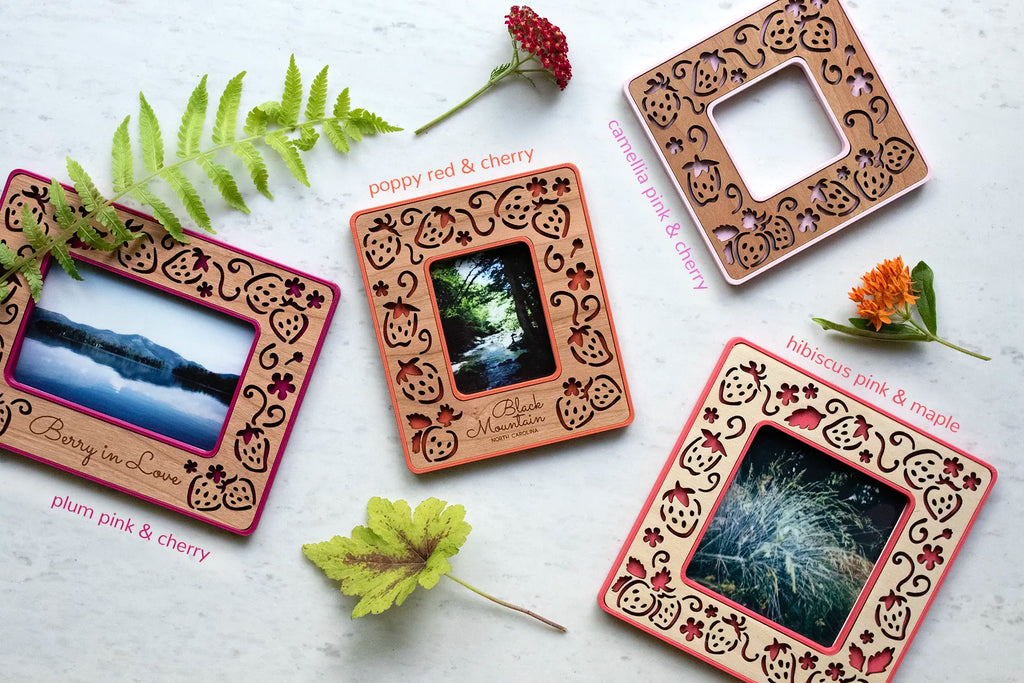 An assortment of colorful, laser cut strawberry Polaroid frames. The frames are lying on a white surface, surrounded by flowers, fern fronds, and leaves. These magnetic frames have an intricate, laser cut strawberry design with real wooden inlays.