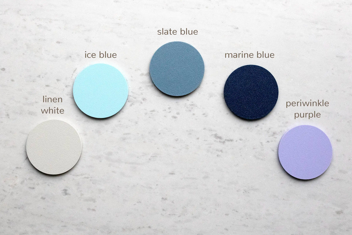 Five 3D-printed color samples for the snowflake picture frame housing, arranged on white marble and labeled: linen white, ice blue, slate blue, marine blue, and periwinkle purple. These are the housing color options for the laser-cut snowflake picture frames.