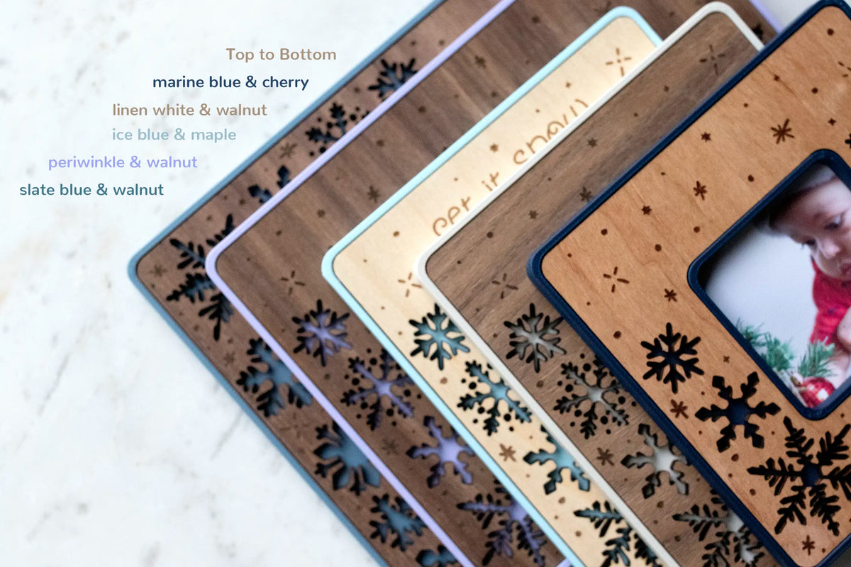 Five laser-cut snowflake magnetic frames fanned out to show the color and wood options — marine blue, linen white, ice blue, periwinkle, and slate blue, in cherry, walnut, and maple wood inlay. One is engraved "Let It Snow." These small winter keepsake frames stick to the fridge or any magnetic surface and can be personalized with a message or left text-free for an all-snowflake design.