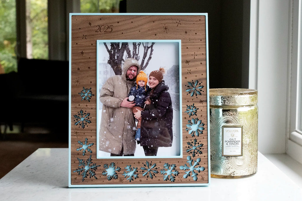 Walnut wood picture frame with laser-cut snowflakes over an ice blue backing, engraved falling snow and "2025," holding a family winter photo. Personalized for a holiday keepsake.