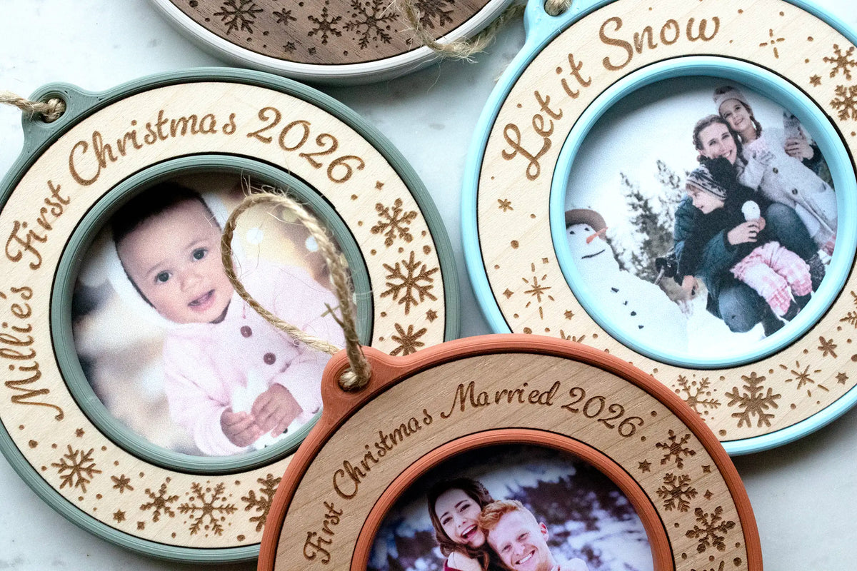 Close-up of three personalized photo ornaments: a sage green and maple baby's first Christmas ornament engraved "Millie's First Christmas 2026," an ice blue and maple "Let it Snow" snowflake ornament, and a terracotta and cherry first Christmas married ornament. Each mini picture frame holds a family photo.