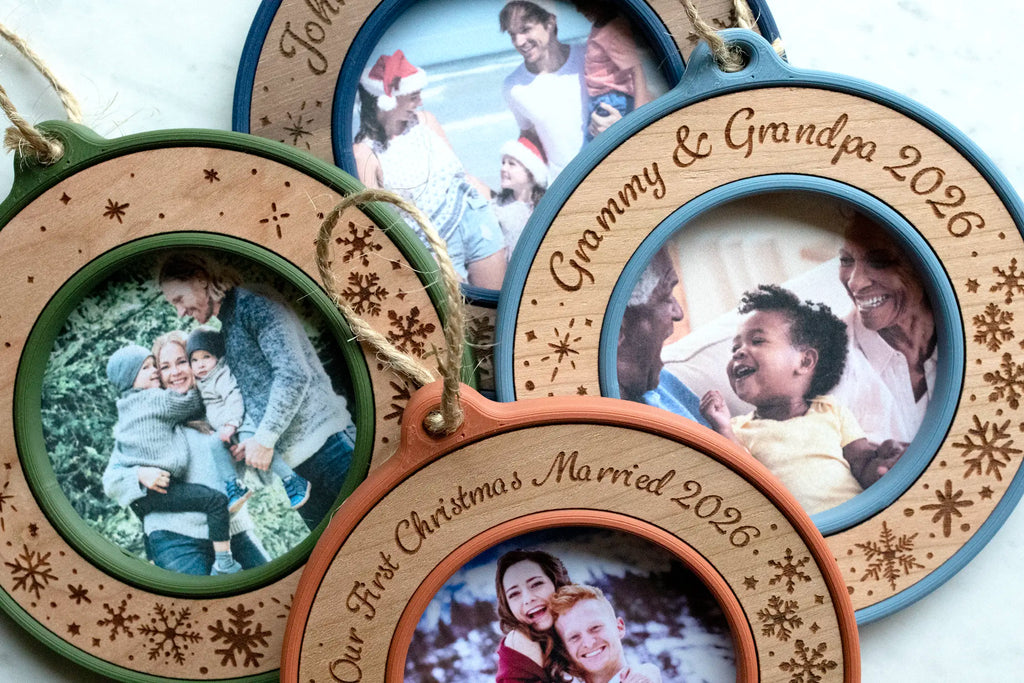 Four personalized Christmas photo ornaments in moss green, marine blue, slate blue, and terracotta, each a mini picture frame with a round photo window and a laser-engraved wood face in cherry or walnut, featuring snowflakes and custom text such as "Grammy & Grandpa 2026" and "Our First Christmas Married 2026." They hold family photos, including grandparents with a baby and a newlywed couple, and hang from jute twine loops.