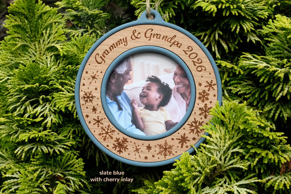 Grandparents Christmas ornament in slate blue with a cherry inlay, laser-engraved "Grammy & Grandpa 2026" with snowflakes, holding a photo of grandparents laughing with their toddler grandchild, hanging on evergreen branches.
