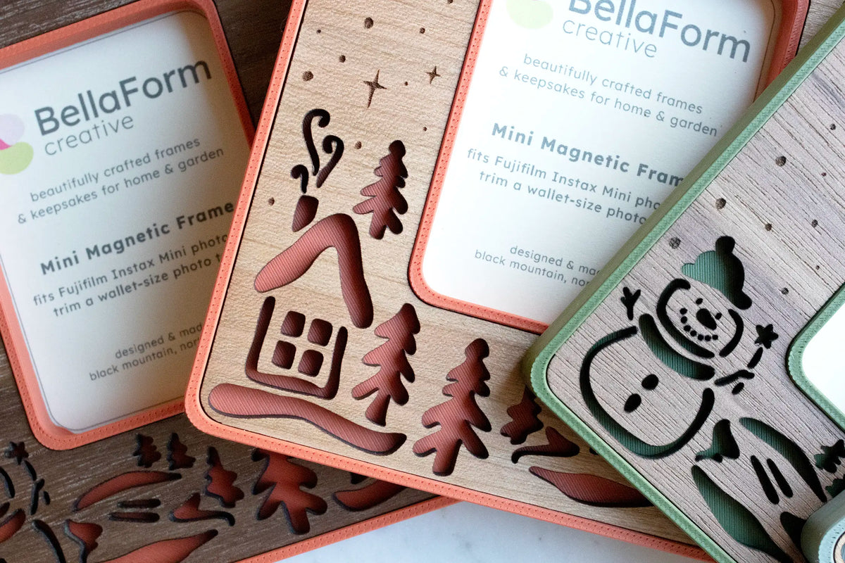 Close-up of snowman magnetic frames showing a laser-cut cabin and evergreen trees over poppy red, and an engraved snowman with falling snow over moss green and walnut.