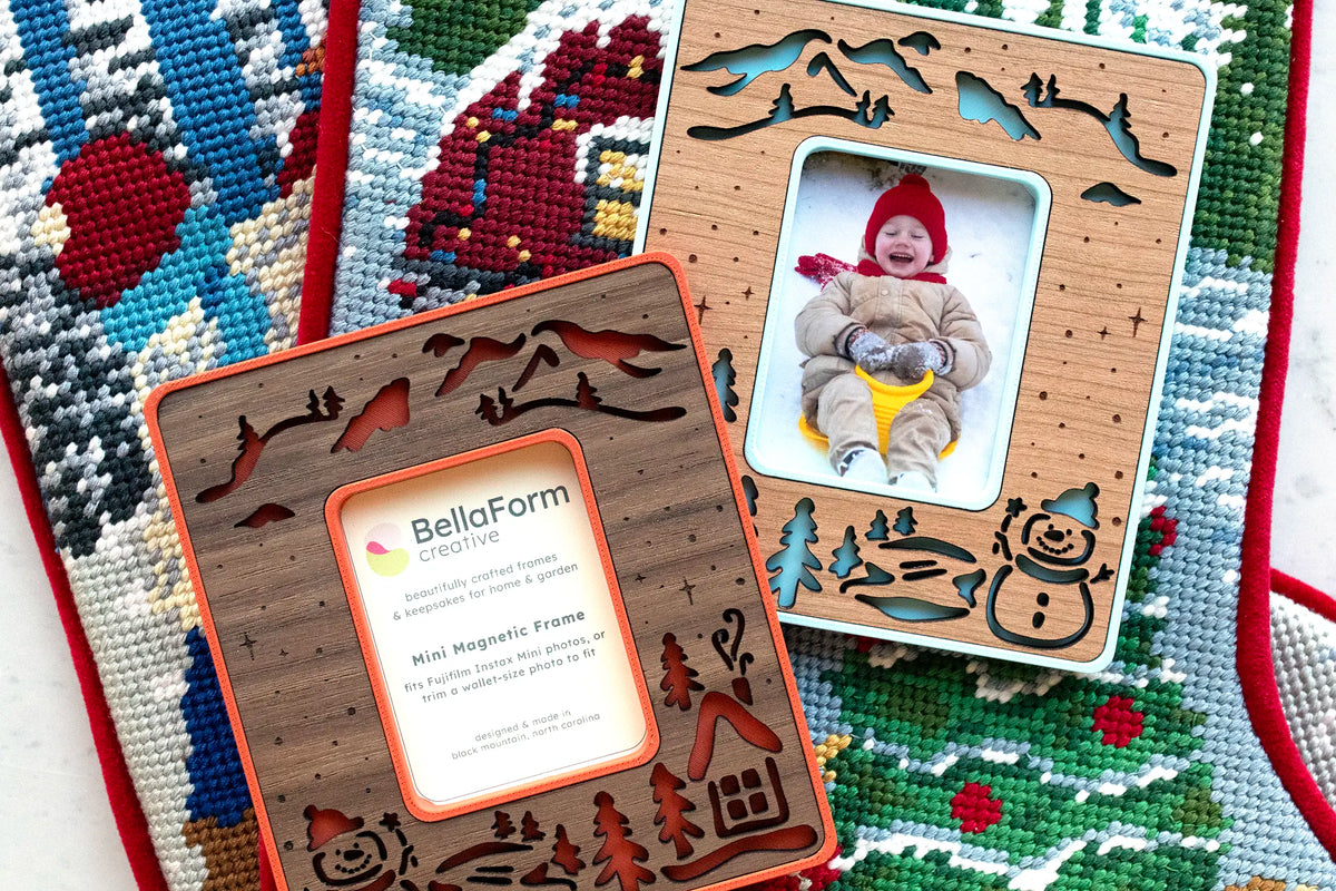 Two snowman magnetic photo frames, one in poppy red and walnut and one in ice blue and cherry holding a photo of a laughing toddler on a sled, resting on a needlepoint Christmas stocking.