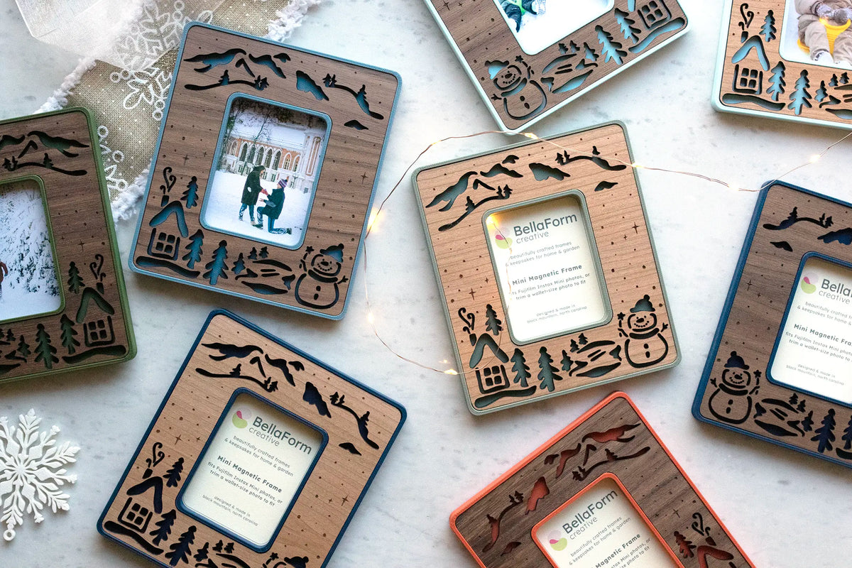Overhead view of snowman Instax Mini magnetic frames in sage green, marine blue, slate blue, ice blue, moss green, and poppy red with walnut and cherry wood inlays, laid out with string lights and snowflakes.