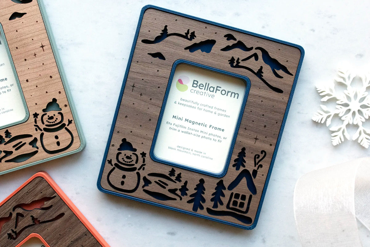 Marine blue snowman magnetic frame with a laser-cut walnut wood inlay of a winter scene with a snowman, cabin, pine trees, and mountains, showing the BellaForm Creative card that fits Fujifilm Instax Mini and wallet-size photos.