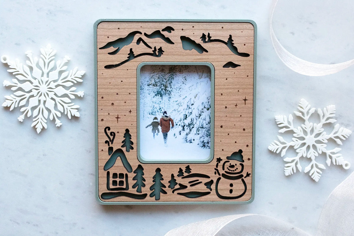 Snowman magnetic picture frame in sage green with a laser-cut cherry wood inlay of mountains, a cabin, pine trees, a frozen pond, and a snowman, holding a photo of two children running through a snowy forest, styled with white snowflake ornaments.