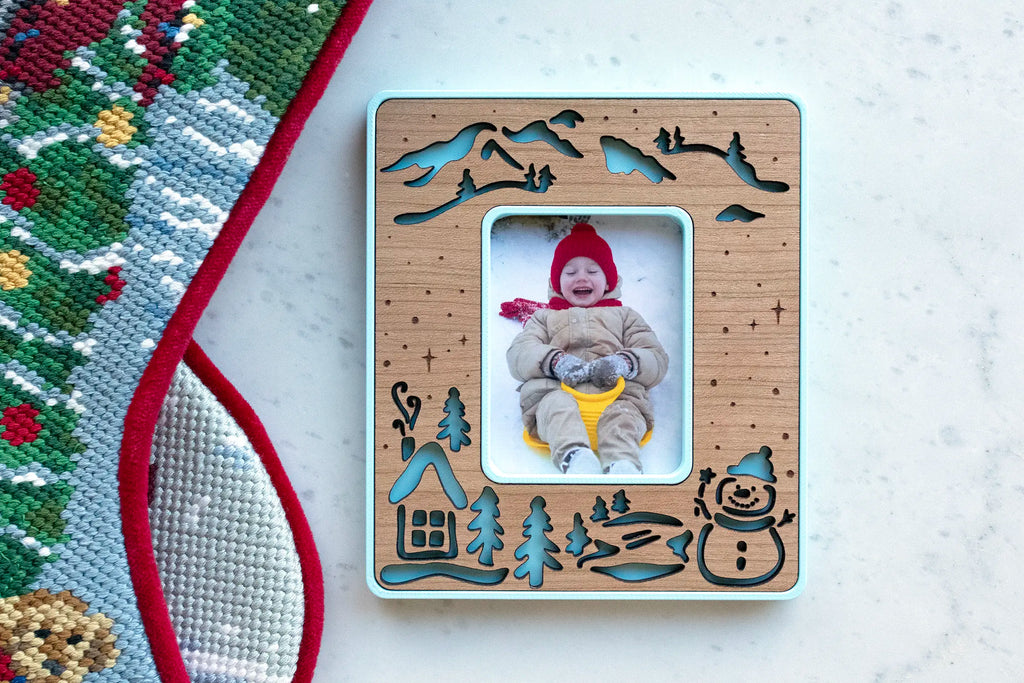 Ice blue snowman magnetic photo frame with a cherry wood inlay of a snowy cabin, pine trees, mountains, and a snowman, holding a photo of a toddler in a red hat sledding, beside a Christmas stocking.